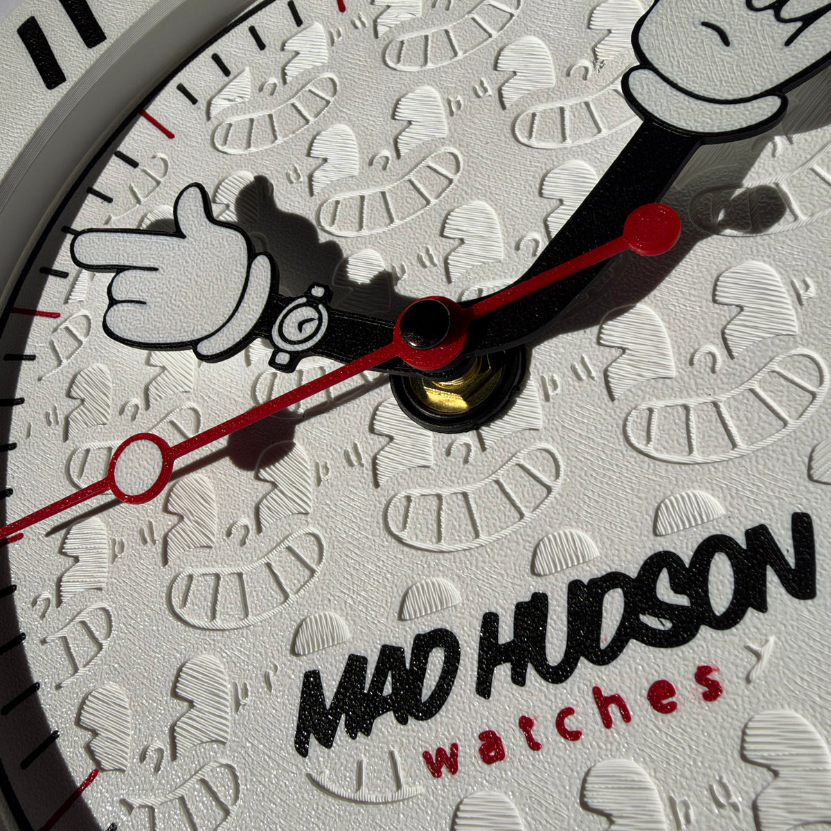 Close-up of a white watch dial featuring an embossed peanut pattern, black cartoon hand graphics, red hands and markers, and the text 'MAD HUDSON watches'.