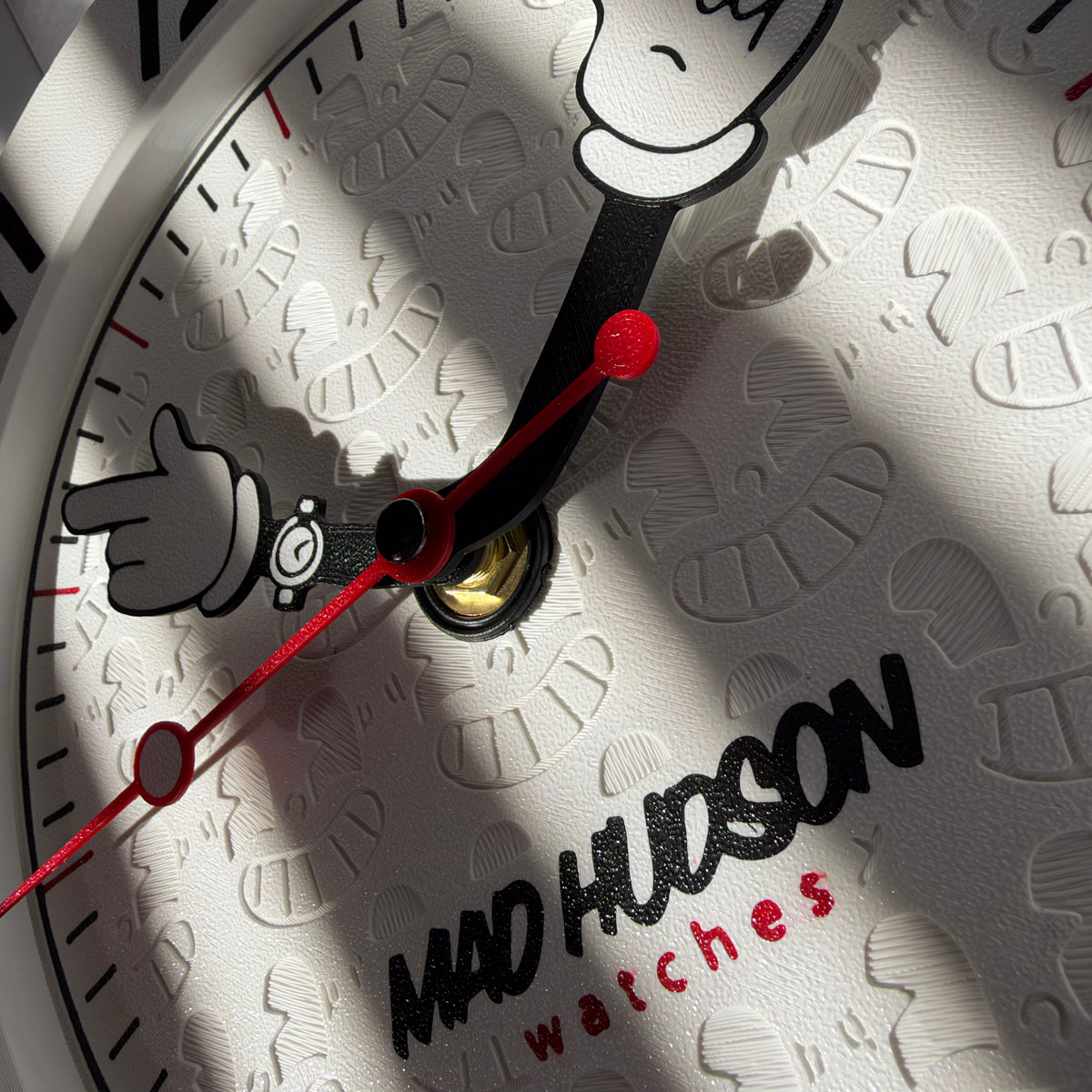 Close-up of a white watch dial with embossed cartoon-style hand shapes, black hands, red accents, and the text 'MAD HUDSON watches' near the bottom.
