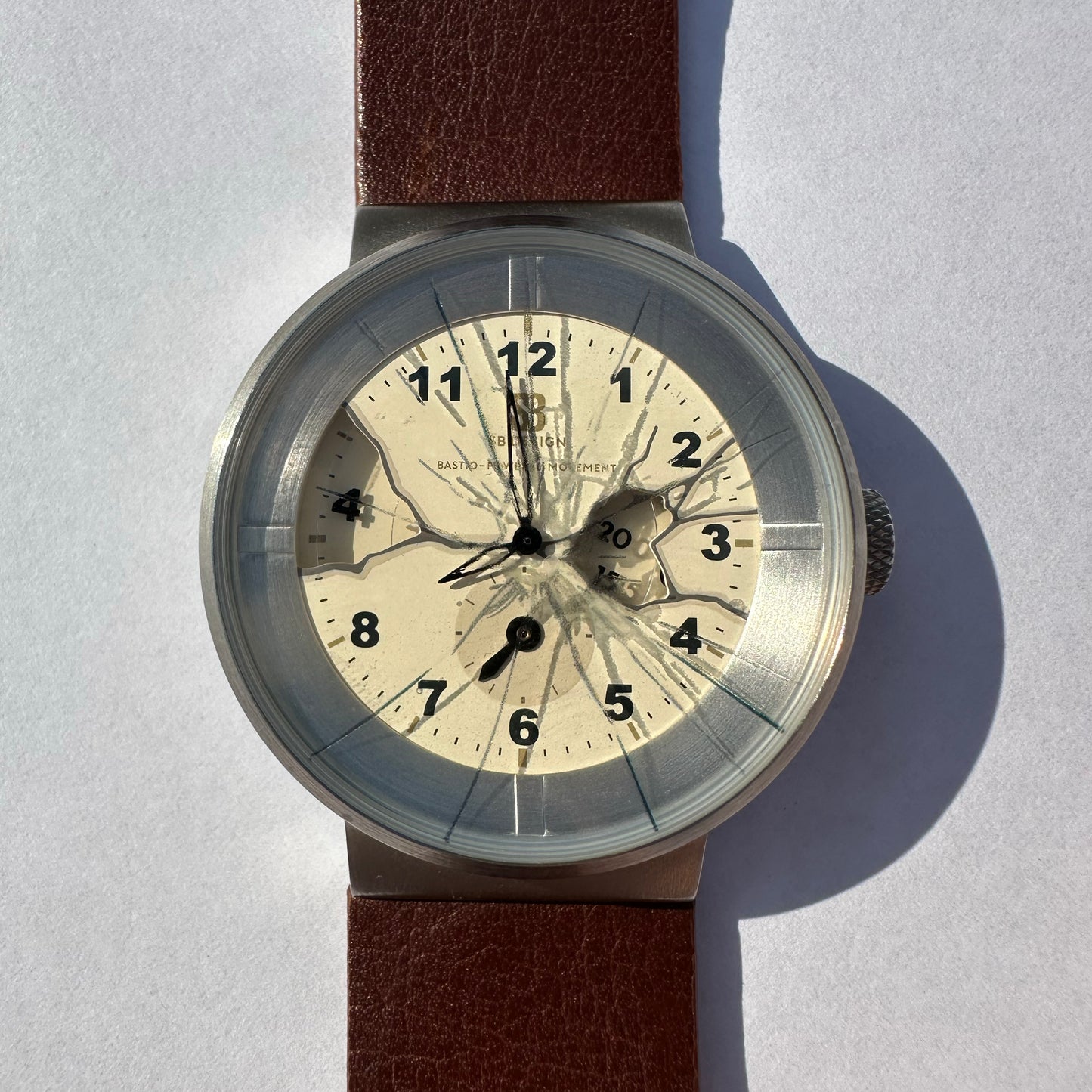 A vintage-style wristwatch with a round silver-toned case, a cream-colored dial featuring a cracked-glass motif and black numerals, and a brown leather strap, photographed on a light gray background with a hard shadow.