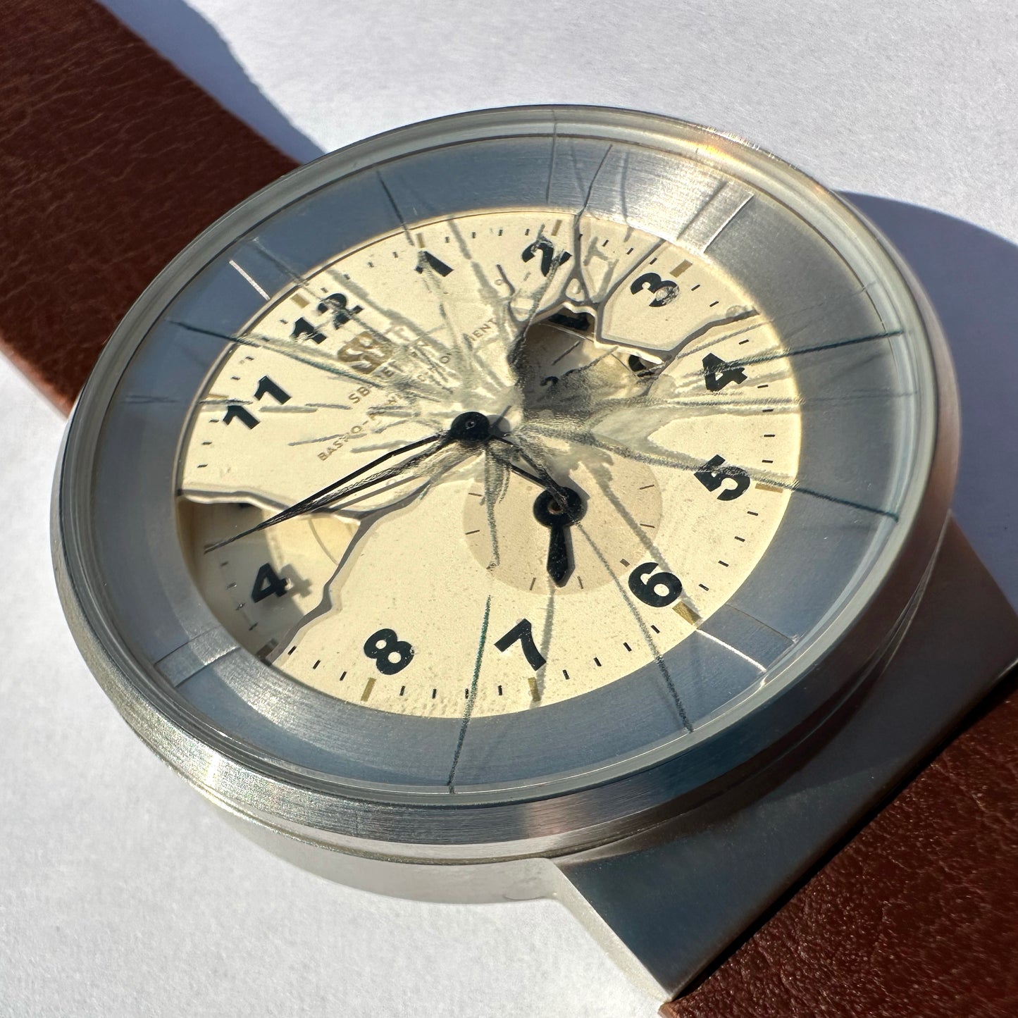 A close-up of a vintage-style wristwatch with a cracked clear crystal, cream-colored dial, black Arabic numerals, and a silver-toned case. A brown leather strap is partially visible on a white background.