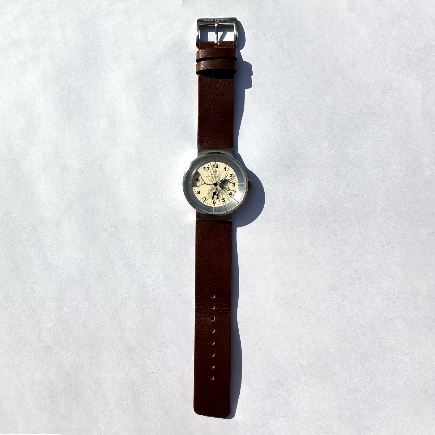 A vintage-style wristwatch with a brown leather strap, silver-toned metal case, and a cream-colored dial, displayed vertically on a light gray background.
