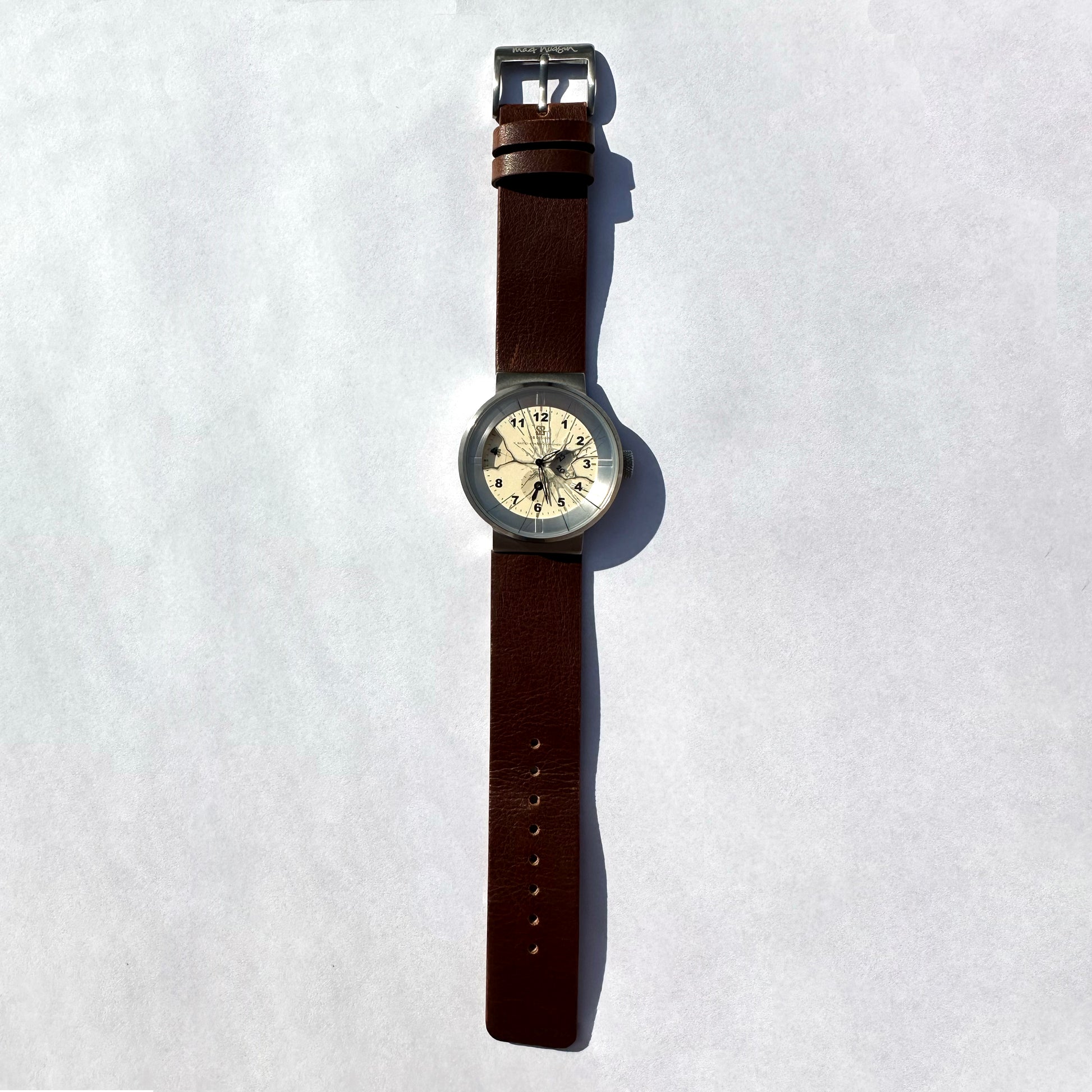 A vintage-style wristwatch with a brown leather strap, silver-toned metal case, and a cream-colored dial, displayed vertically on a light gray background.
