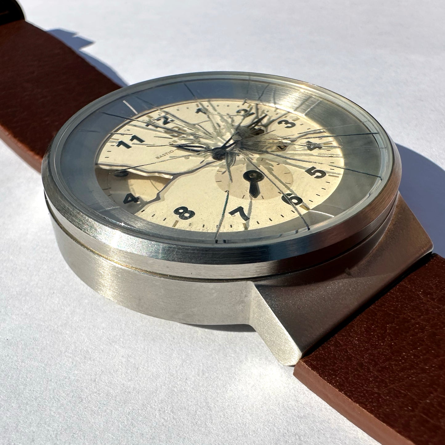 A close-up of a wristwatch with a brushed silver-tone metal case, a cream-colored dial, and a brown leather strap on a white textured background.