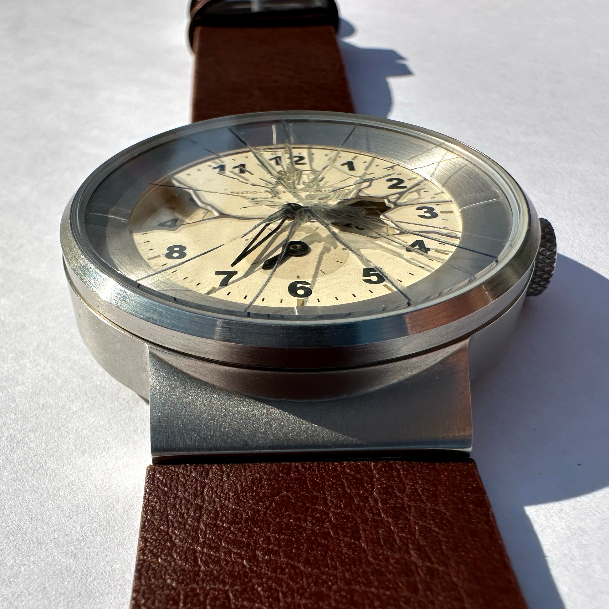 Vintage-style wristwatch with a round silver-toned case, cream dial with black numerals, and a brown leather strap, photographed on a white surface in bright sunlight.