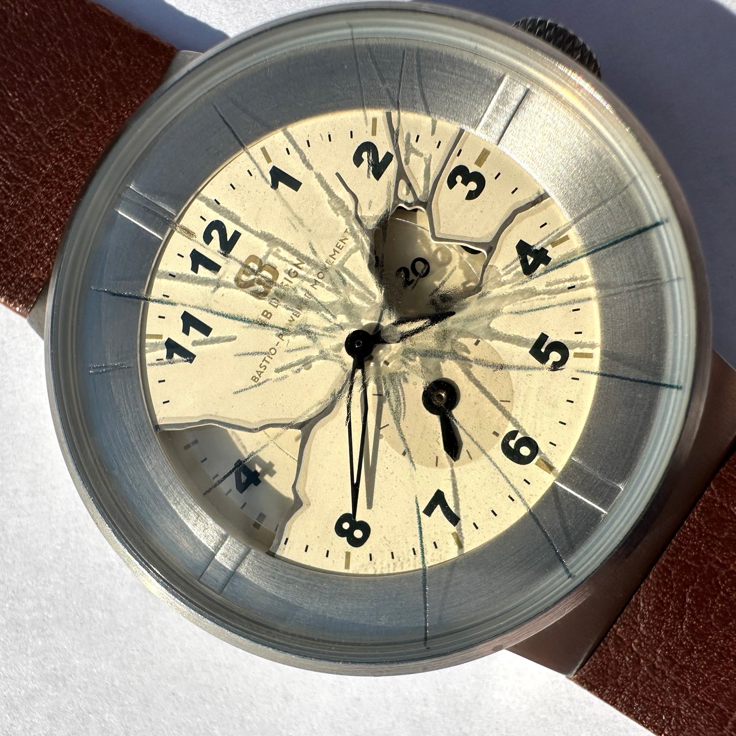 Close-up of a vintage-style wristwatch with a round silver-toned case, cream dial, black Arabic numerals, and a cracked transparent dial overlay. A brown leather strap is partially visible at the top left and bottom right.