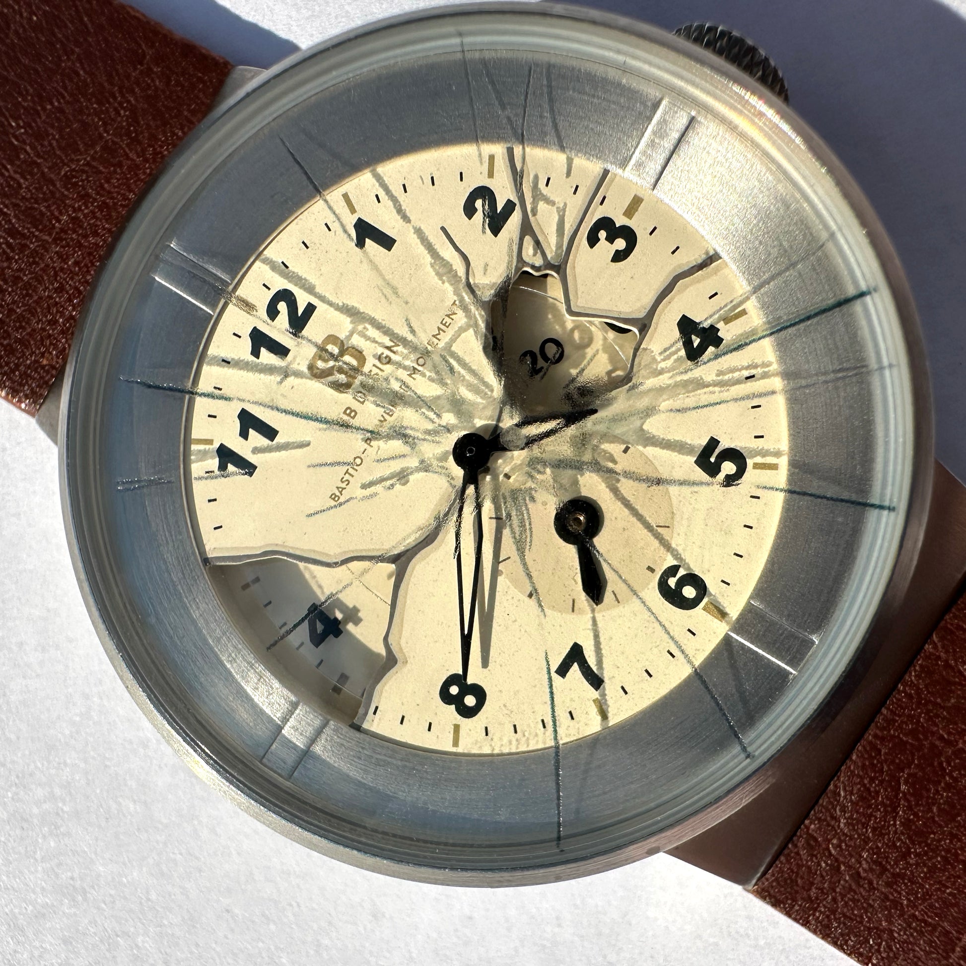 Close-up of a vintage-style wristwatch with a round silver-toned case, cream dial, black Arabic numerals, and a cracked transparent dial overlay. A brown leather strap is partially visible at the top left and bottom right.