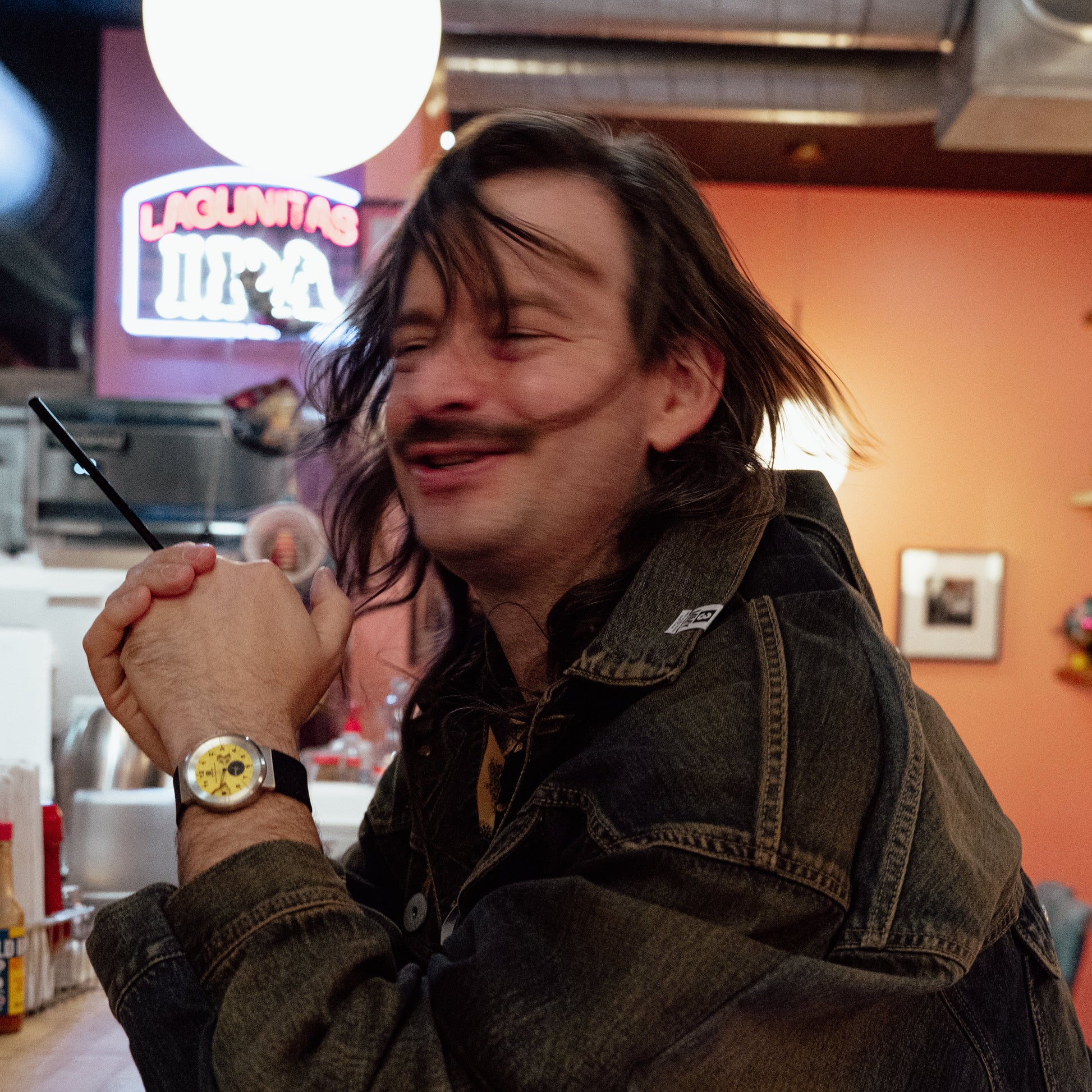 Man with a mustache wearing a watch in a casual diner with neon signs and shelves.  Brad Podray Designer of the Broken Time Watch