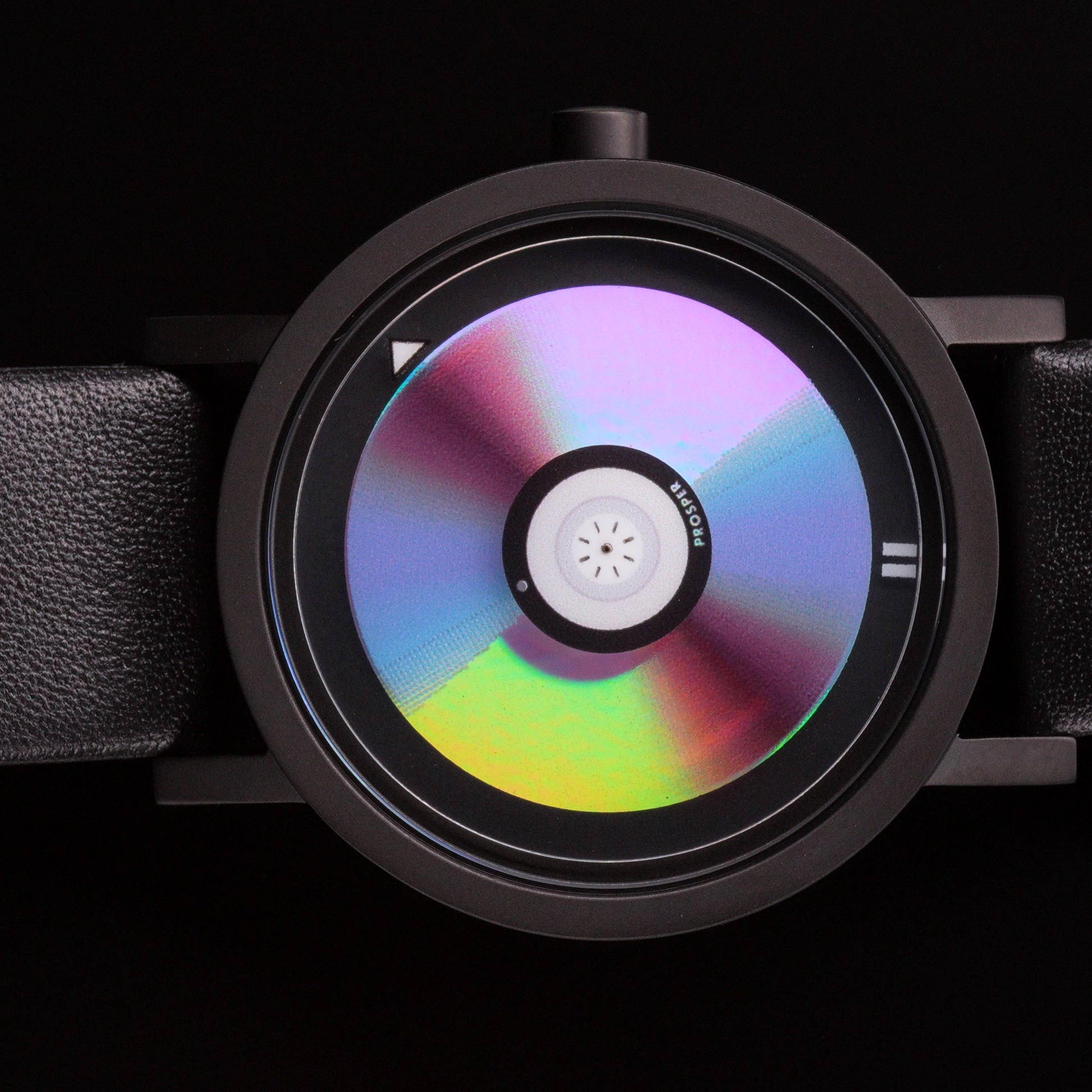Wristwatch on its side that looks like a CD with a blue red and iridescent face with a black stainless steel case and black leather strap on a black background CD Watch Riki Prosper Mad Hudson