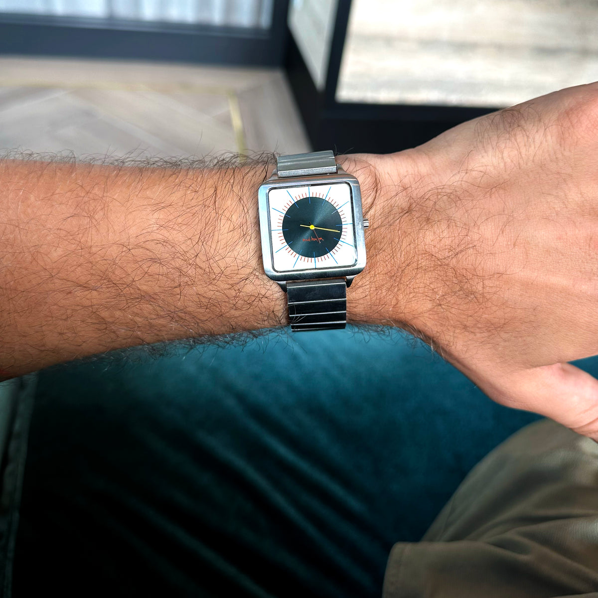 A square analog watch with a silver-toned metal case and link bracelet, featuring a black dial with a yellow second hand, worn on a person's wrist.