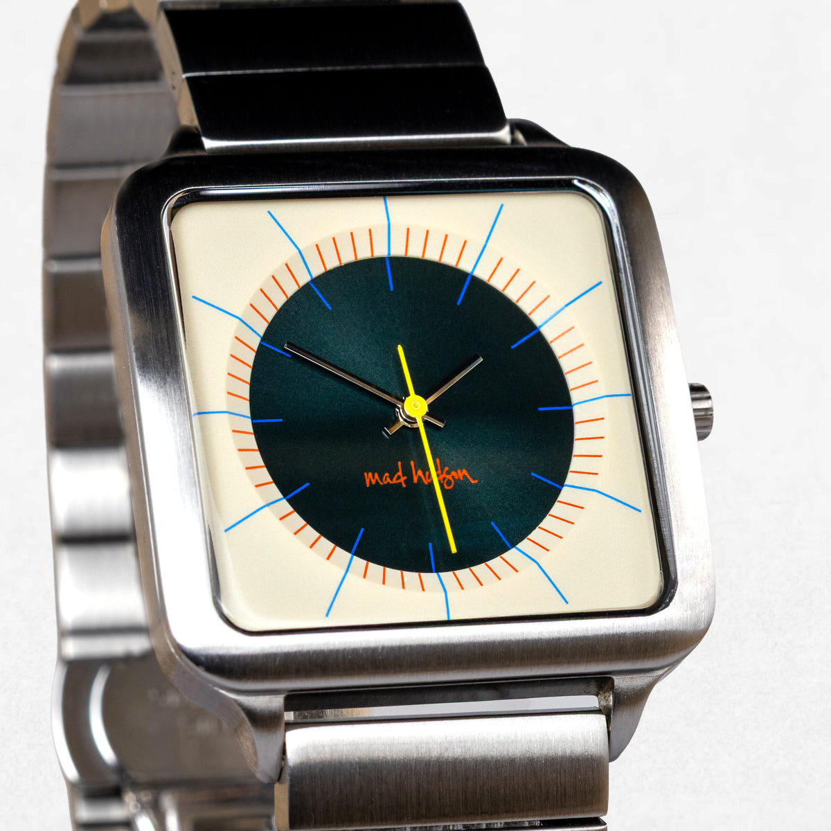 A square analog watch with a brushed silver metal case and link bracelet, featuring a cream-colored dial with a dark green center, orange and blue markings, and a yellow second hand. The brand name 'mad hagem' is printed in orange on the dial.