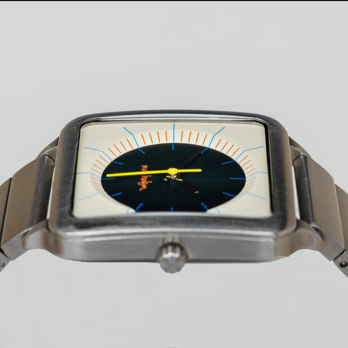 A square analog wristwatch with a brushed silver-tone metal case and bracelet, featuring a cream dial with a large black central section, orange and blue markings, and a yellow hour hand.