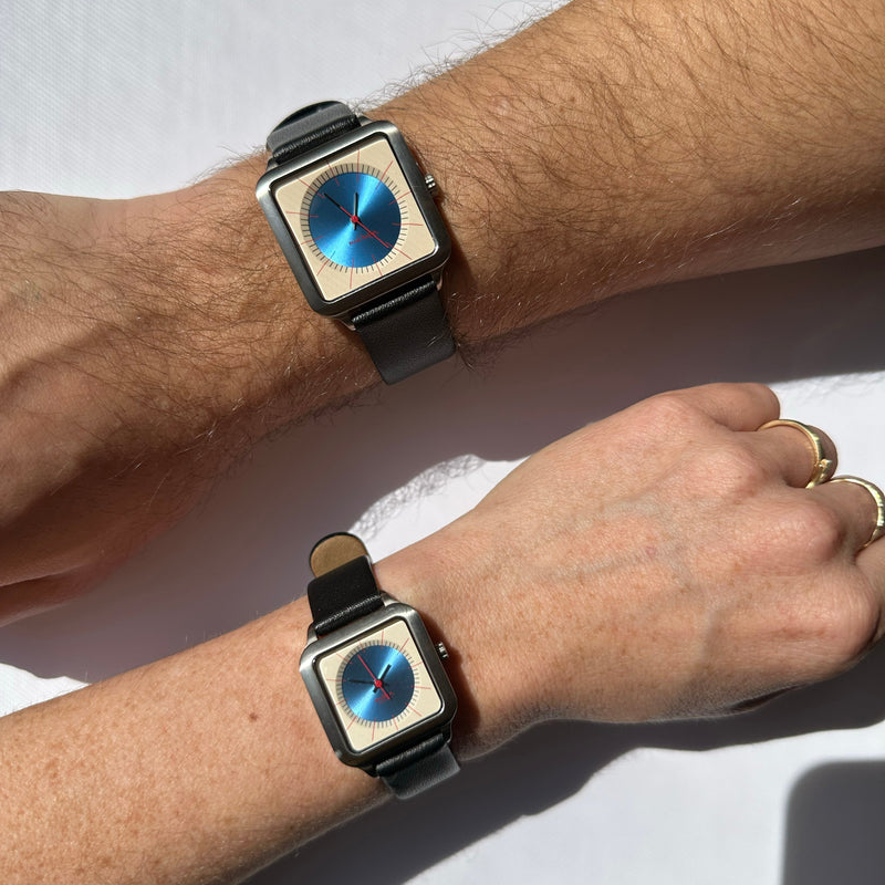 Two square watches with black straps and metallic silver-toned cases, each featuring a blue circular dial, worn on human wrists against a light background.