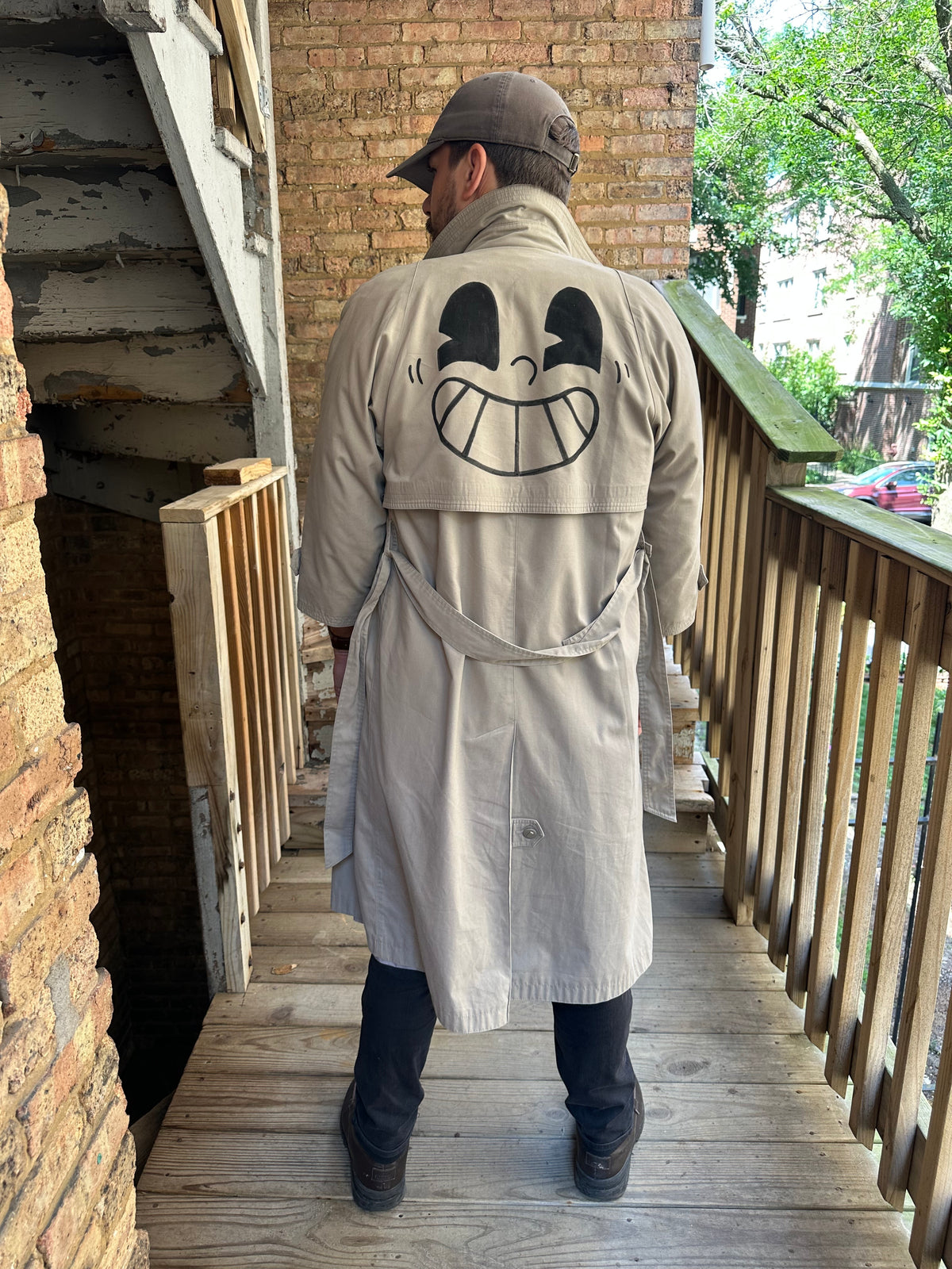 A man viewed from behind wearing a beige trench coat with a large black cartoon face graphic printed on the back, standing on a wooden porch.