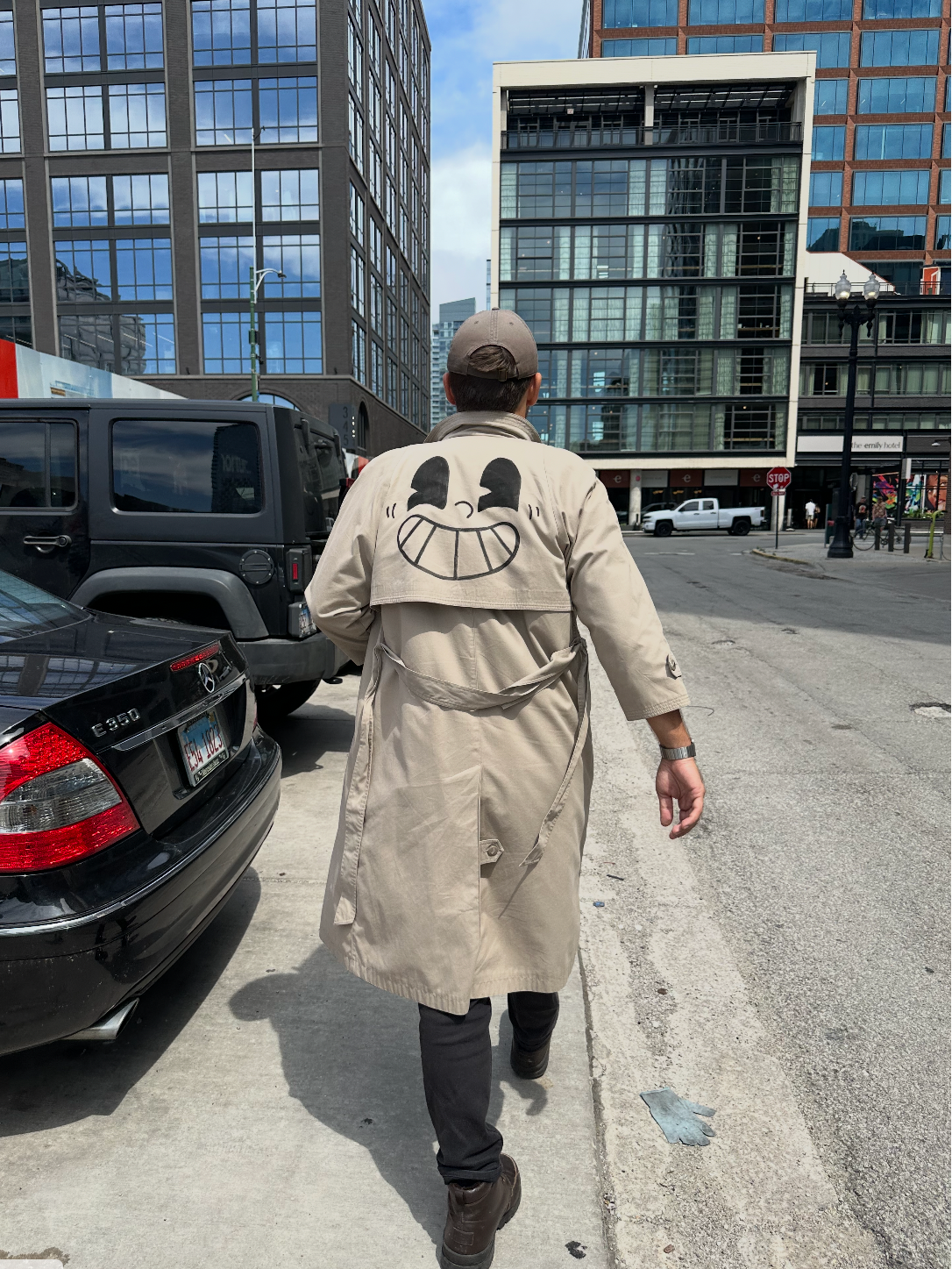 A man walking away on a city street wearing a beige trench coat with a large cartoon smiley face graphic printed on the back, dark pants, and a cap. Parked cars and modern buildings are visible in the background.