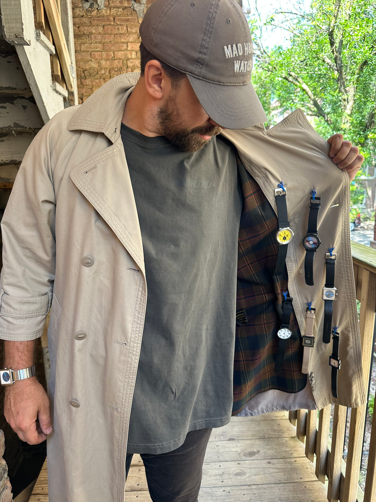 A man wearing a beige trench coat over a dark gray t-shirt, holding the coat open to display a collection of wristwatches hanging on the inside lining. He is wearing a brown baseball cap with white text that reads 'MAD MAX WATCHES'.