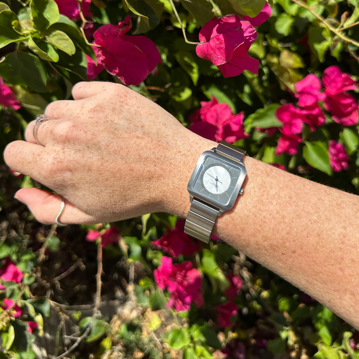 A silver-toned square analog watch with a matching metal link bracelet worn on a person's wrist, photographed outdoors in front of bright pink flowers and green foliage.