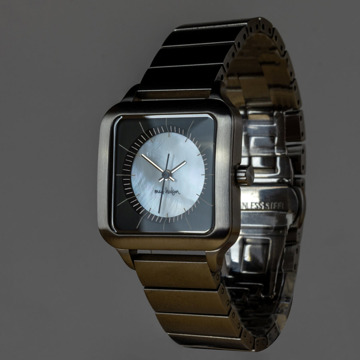A square-faced wristwatch with a dark gunmetal-tone metal case and link bracelet, featuring a white mother-of-pearl dial with slim silver-tone hands. The brand name 'Mossy Oak' is visible on the dial, and 'LESS SIEFF' is engraved on the clasp.