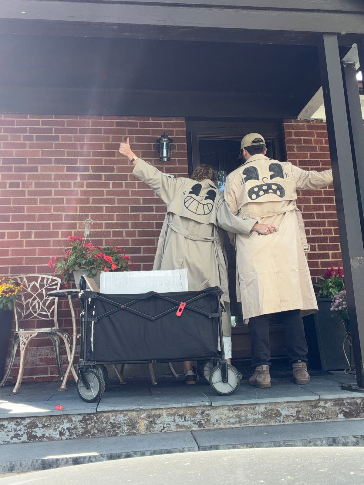 two cute people in trench coats witch cartoon faces drawn on the back in front of a house